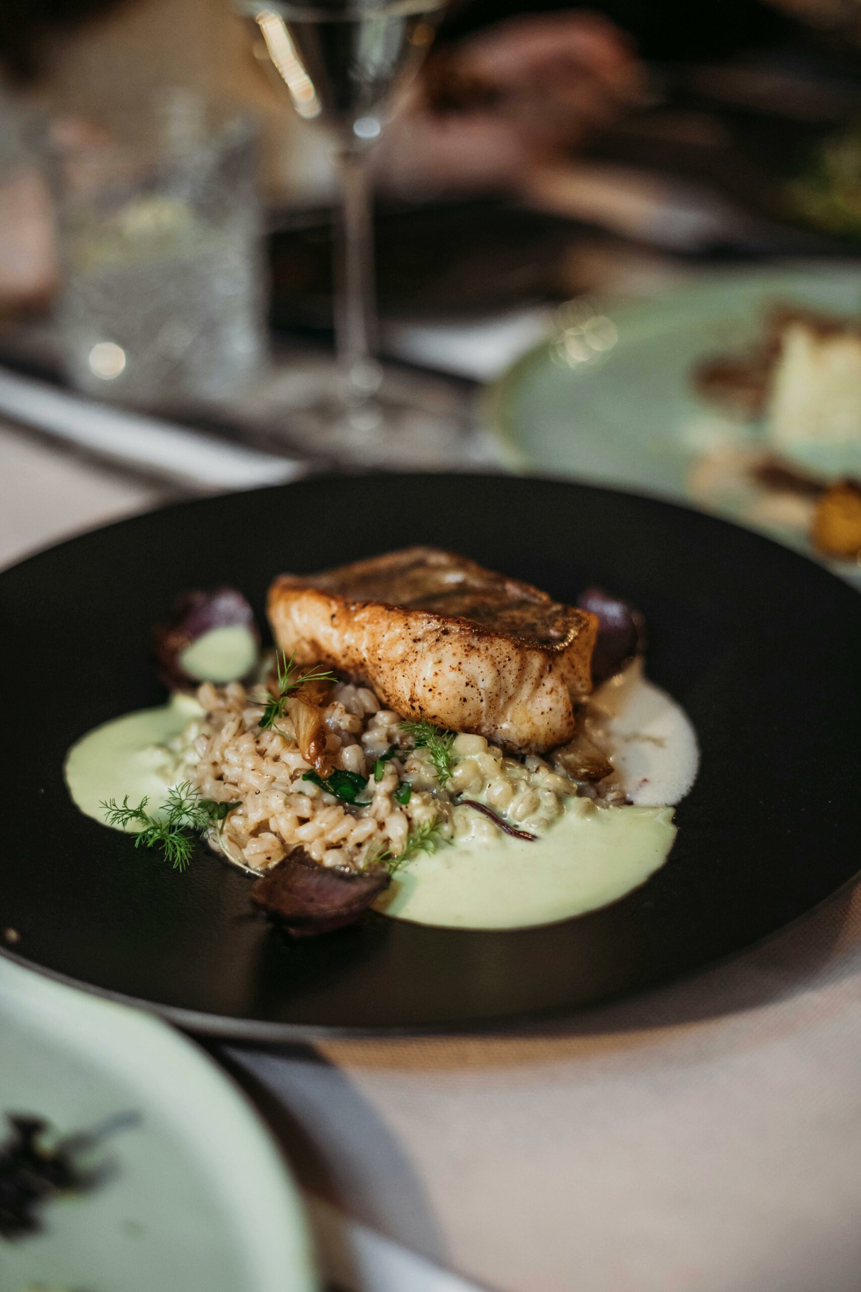 Tasty grilled fish with grains and sauce served on black plate on table with wineglass and dishware in modern light cafe